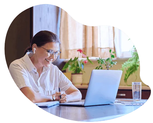 Woman with earbuds smiling during a virtual video call on her laptop, taking notes at a home desk.