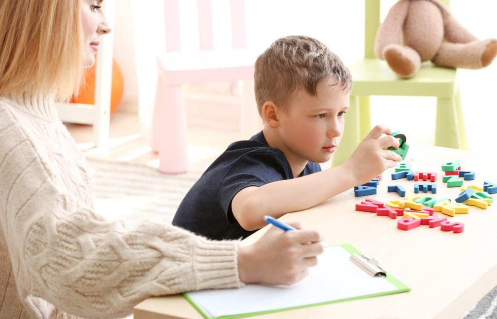 Clinician taking notes on a clipboard while a young boy plays with colorful letter blocks during an evaluation.