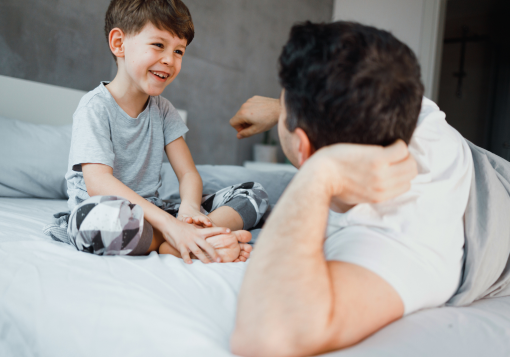 Boy in pajamas laughing while playing a hand-clapping game with his father on a bed.