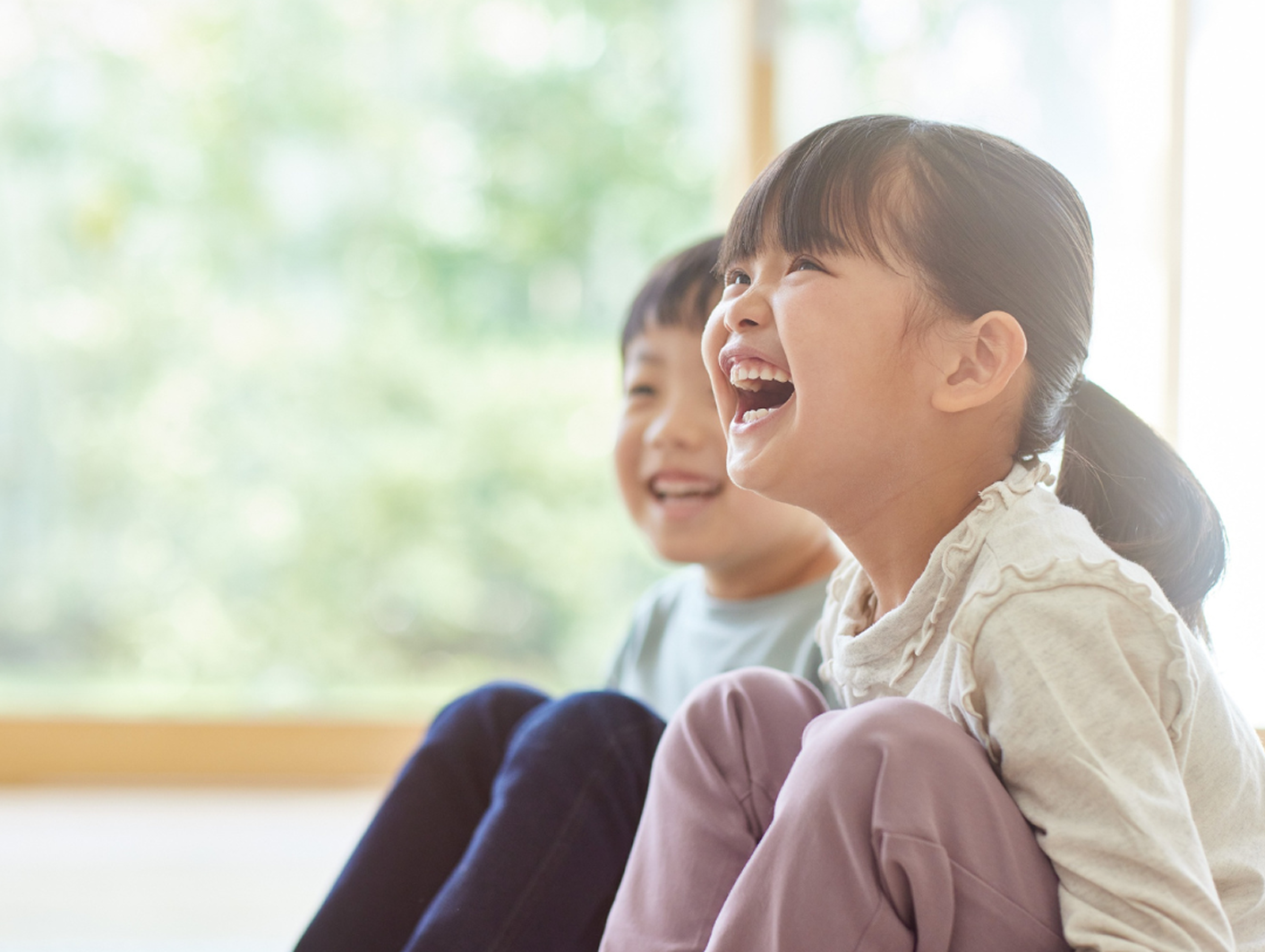 Two young children sitting together and laughing, next to a bright window.