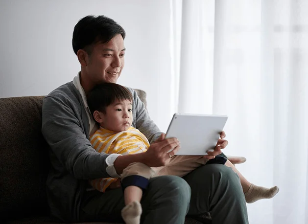 "Father and young son sitting together on a couch, looking at a tablet."