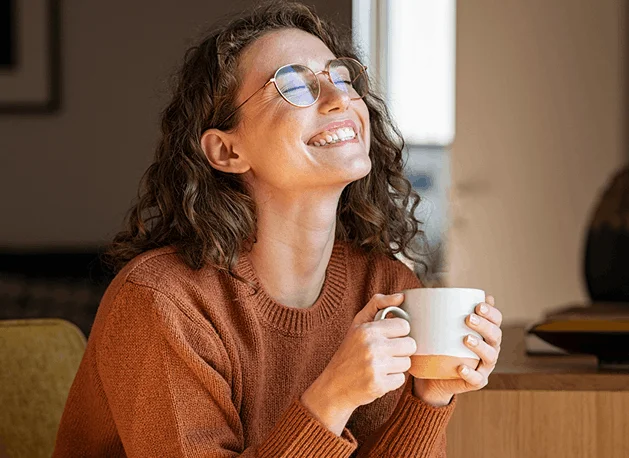 "Woman laughing joyfully while holding a mug, seated by a window."