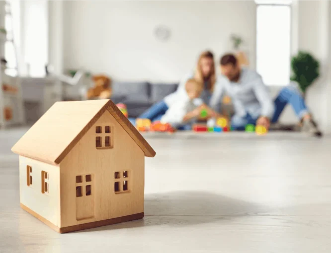 Wooden toy house in the foreground with a family playing together on the floor in the background.