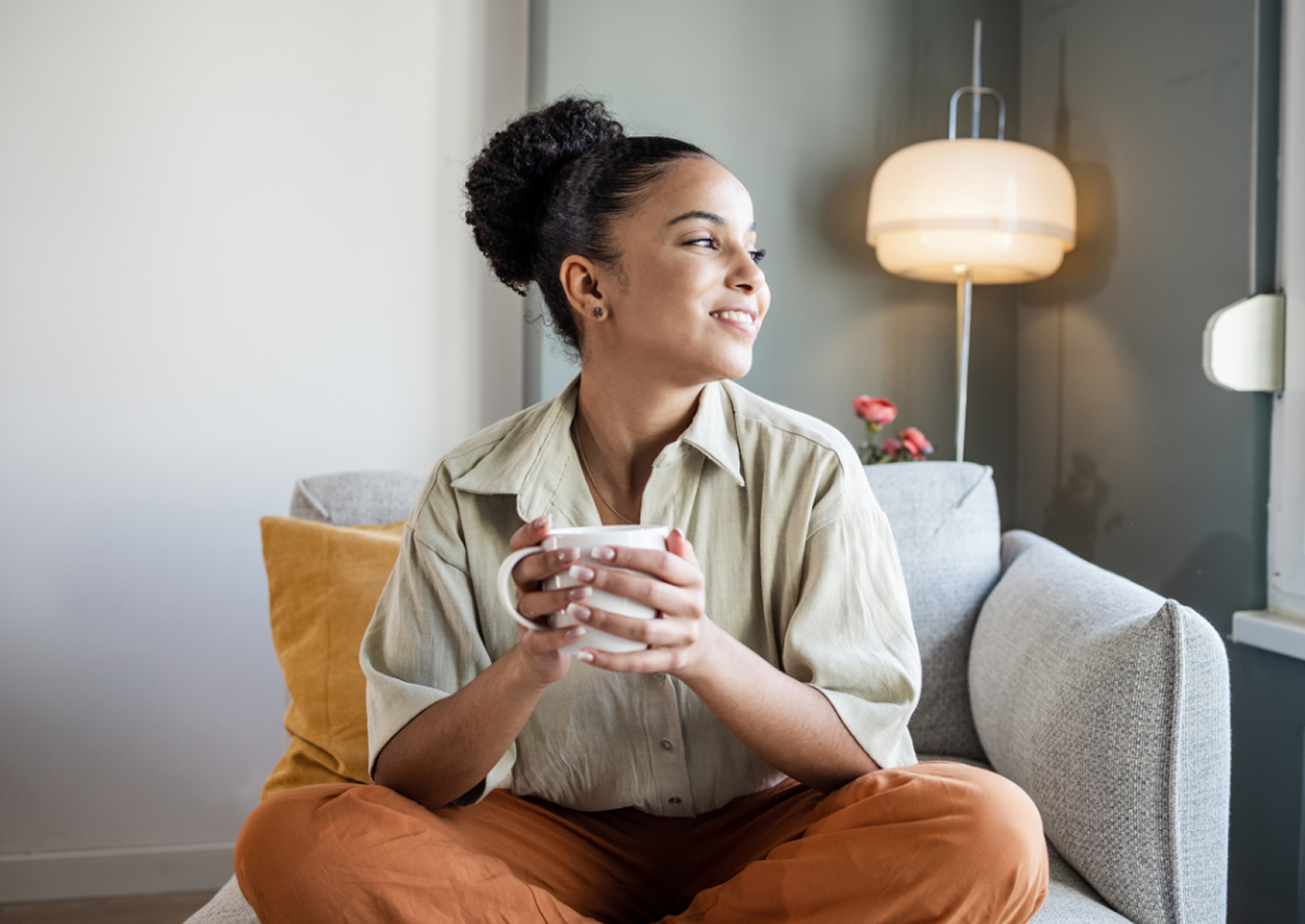 Woman sitting cross-legged on a couch, holding a mug and smiling as she looks out a window.