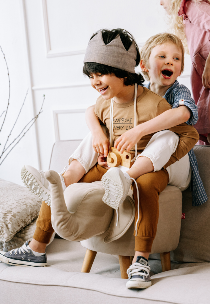Boy in a fabric crown playing with a toy camera while another boy laughs and hugs him from behind.