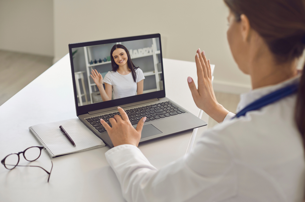 Clinician in a white coat waving during a video call with a smiling patient on a laptop screen.