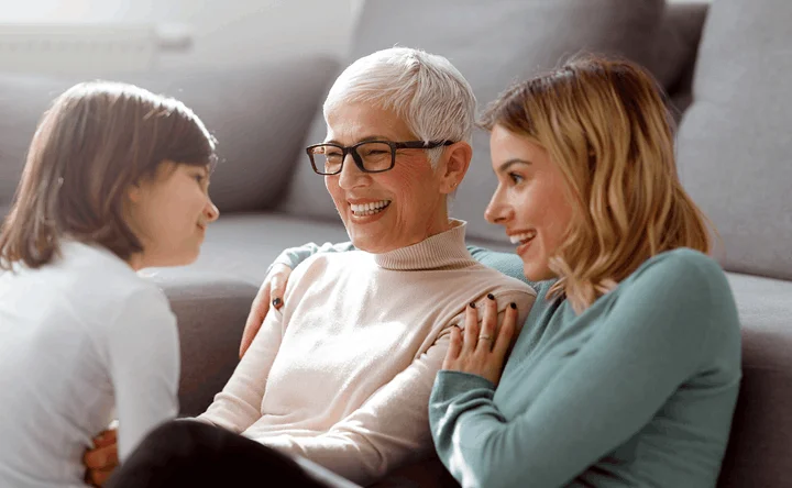 Three generations of a family — child, grandmother, and mother — laughing together on a couch.