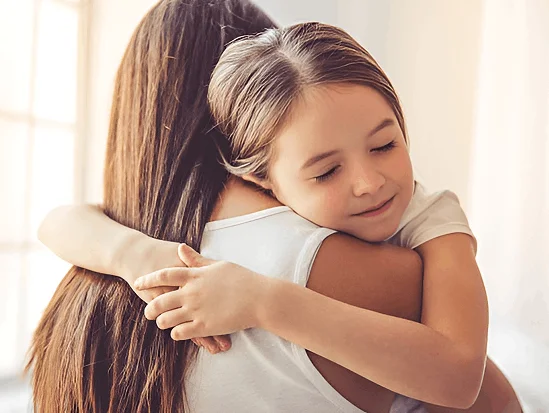 Young girl hugging her mother tightly, resting her chin on her shoulder with eyes closed.